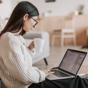 A woman sitting on the floor researching hiring a copywriter on her laptop while drinking from a mug