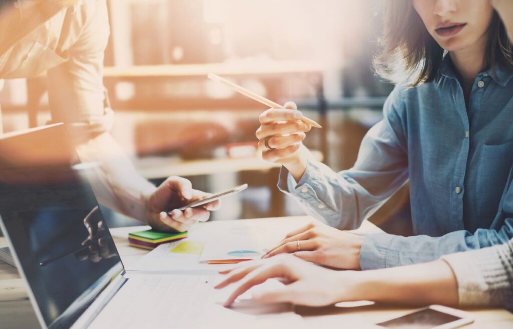 Two people work at a desk with a laptop, documents, and a smartphone, discussing b2b seo strategy while one gestures with a pen.