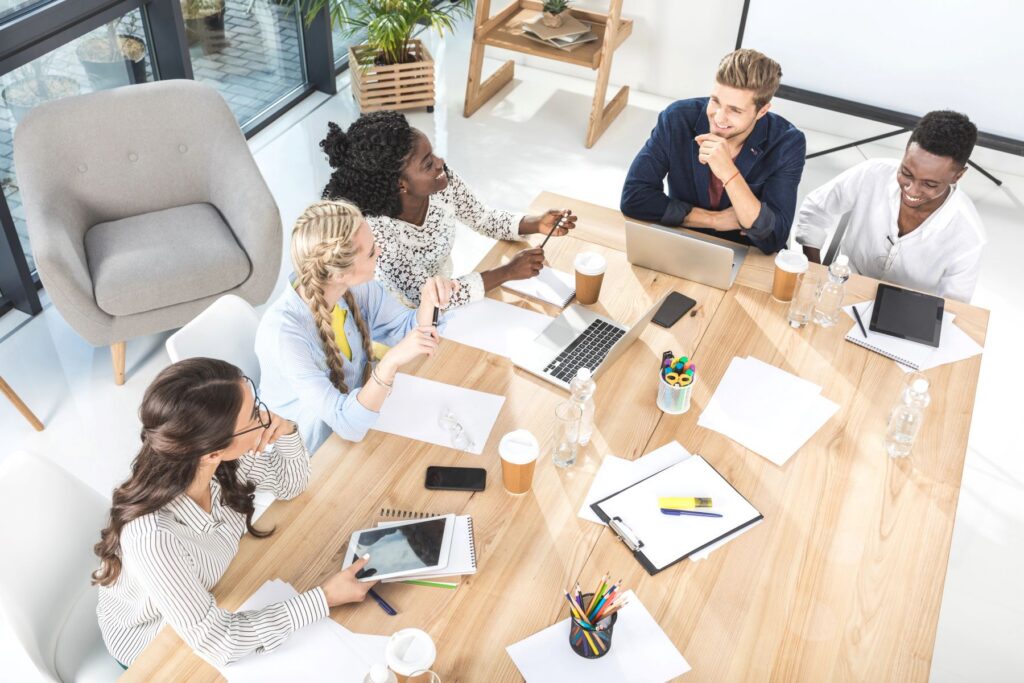 Five people sit around a wooden table in a bright office, with laptops, notebooks, and coffee cups, appearing to discuss work.