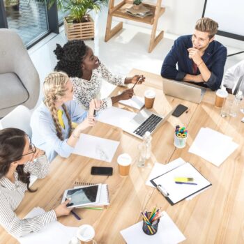 Five people sit around a wooden table in a bright office, with laptops, notebooks, and coffee cups, appearing to discuss work.
