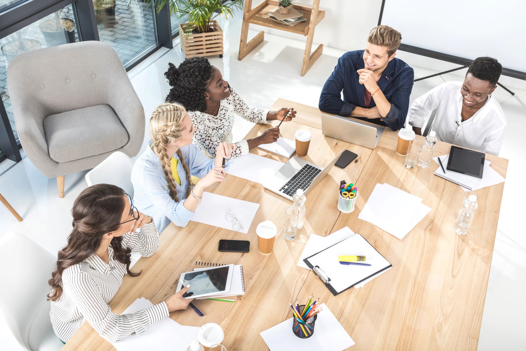 Five people sit around a wooden table in a bright office, with laptops, notebooks, and coffee cups, appearing to discuss work.