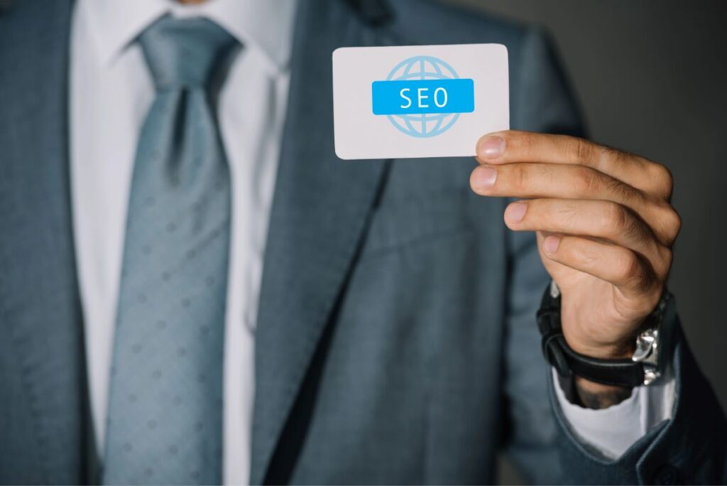 A person in a suit and tie holds a card that displays the word "SEO" in blue letters over a globe icon, symbolizing the importance of an effective B2B SEO strategy in reaching global business audiences.