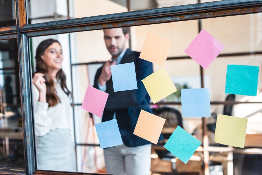 Two people stand behind a glass wall covered with colorful sticky notes, appearing to discuss ideas for a b2b seo strategy in a modern office setting.