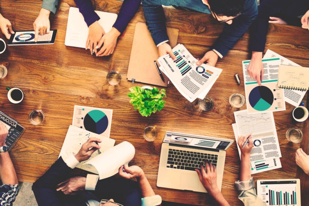 People sit around a wooden table with laptops, documents, and charts, discussing data and business reports in a meeting setting focused on refining their B2B SEO strategy.