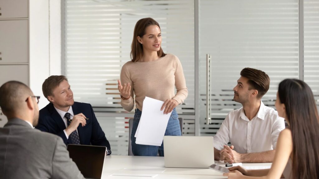 A freelance SEO consultant stands holding papers and speaks to four colleagues seated around a conference table in a modern office setting.