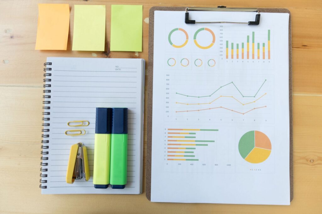 A notepad, highlighters, stapler, paper clips, sticky notes, and a clipboard with charts and graphs are arranged neatly on a wooden desk—essential tools for any freelance SEO consultant.