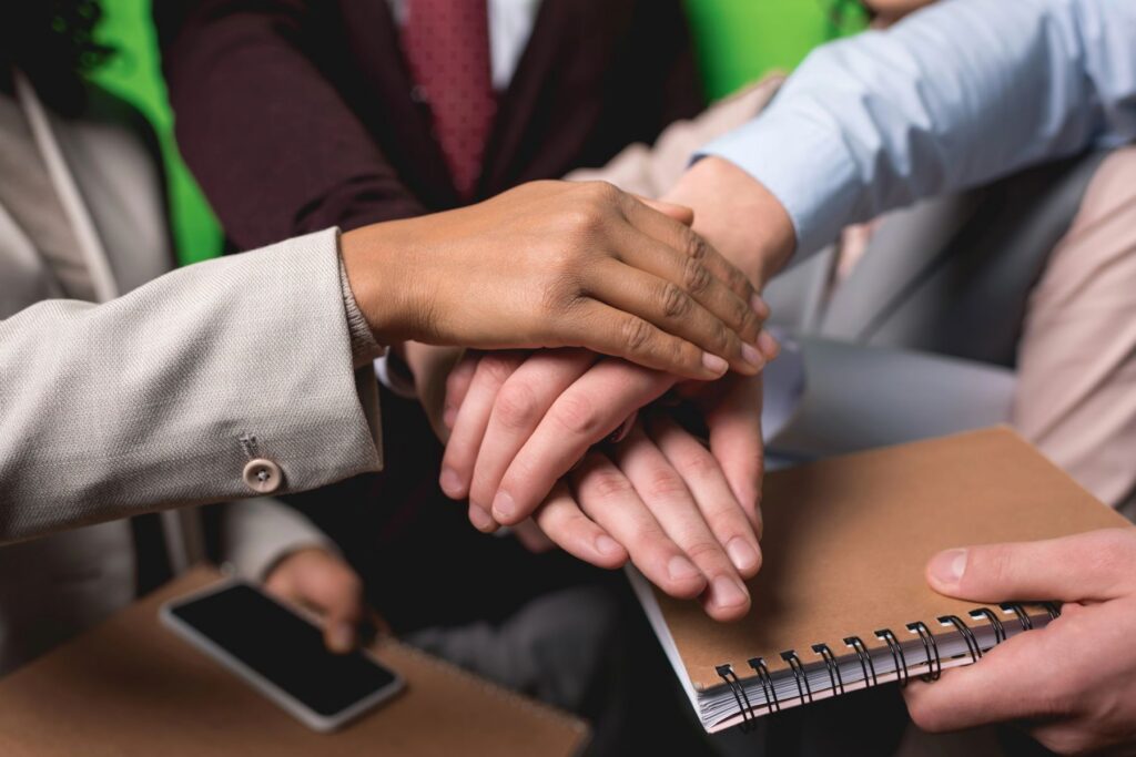 Several people stack their hands in a gesture of teamwork; among them, a freelance SEO consultant holds a notebook while another holds a smartphone.