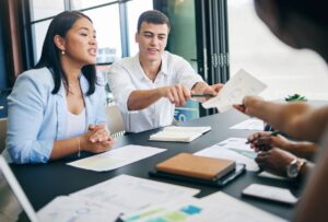 Four people sit at a table with documents, charts, and notebooks, engaged in discussion as a freelance SEO consultant hands a paper to another team member.
