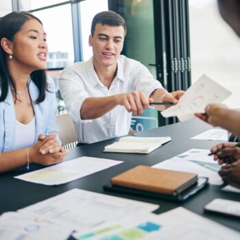 Four people sit at a table with documents, charts, and notebooks, engaged in discussion as a freelance SEO consultant hands a paper to another team member.